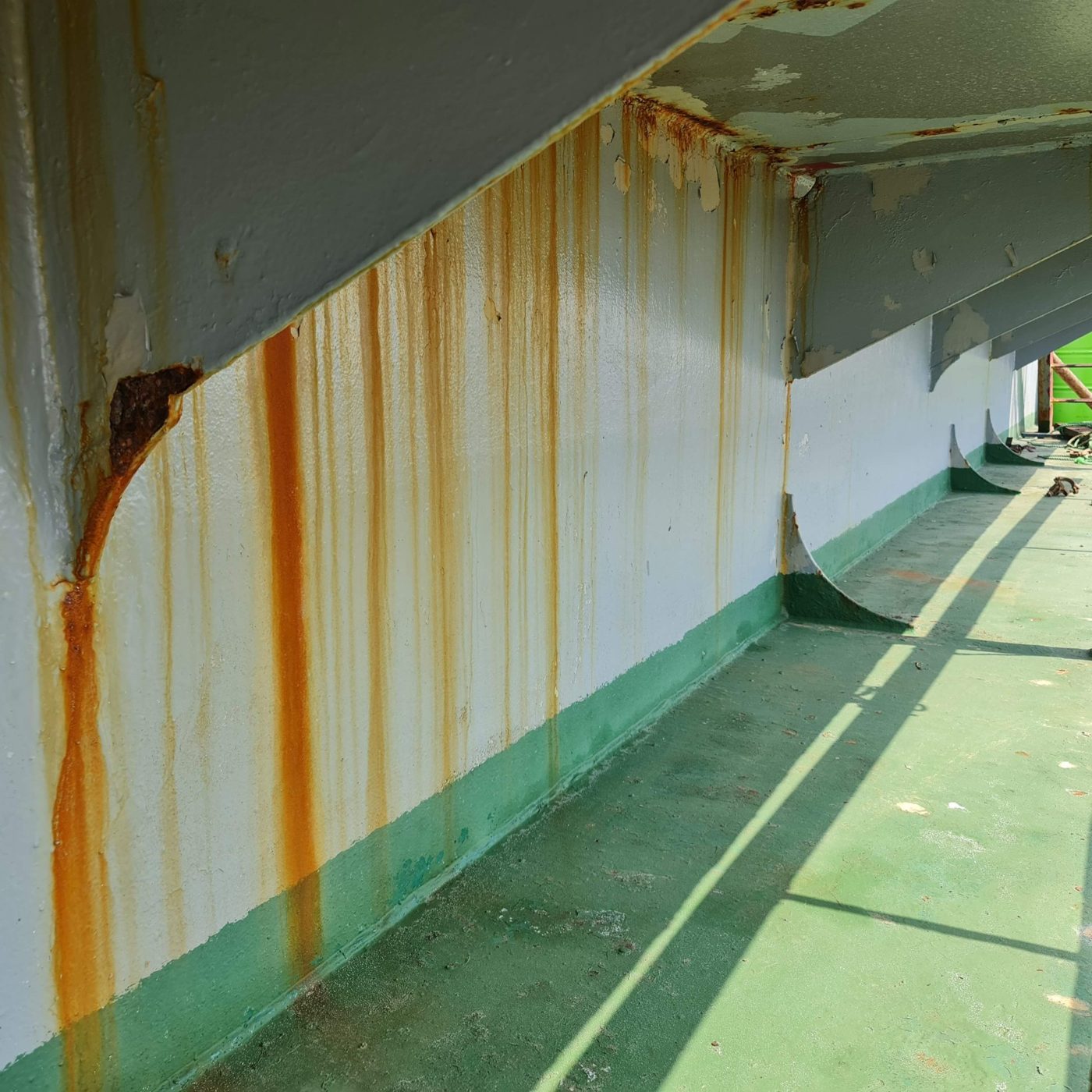 Steel wall with rust streaks running down from a corroded bracket and peeling paint above a green floor coating